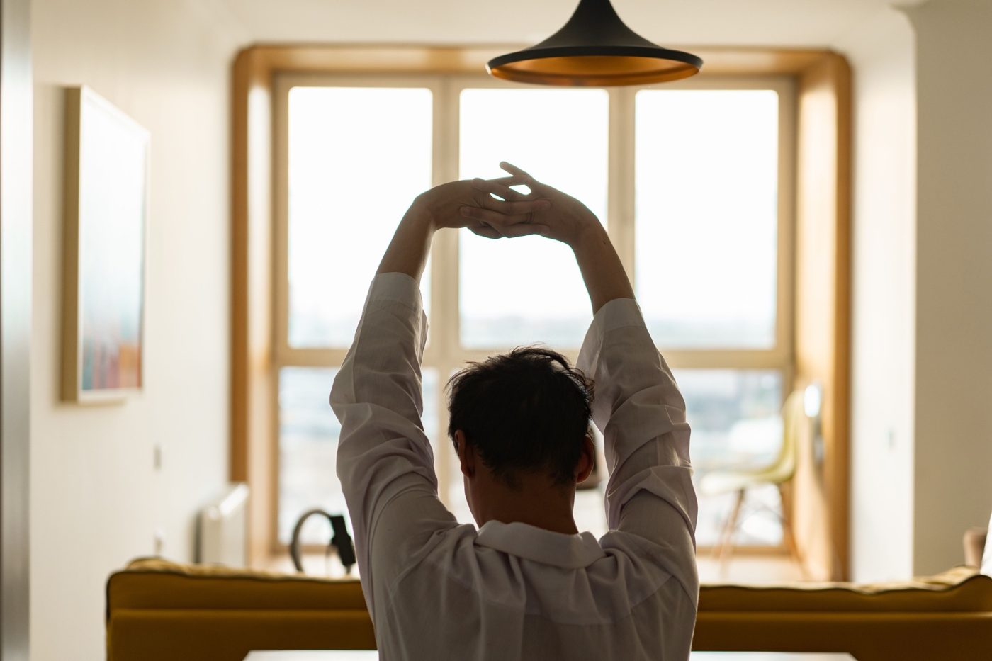 Person stretching with arms raised in front of a bright window at home in the morning