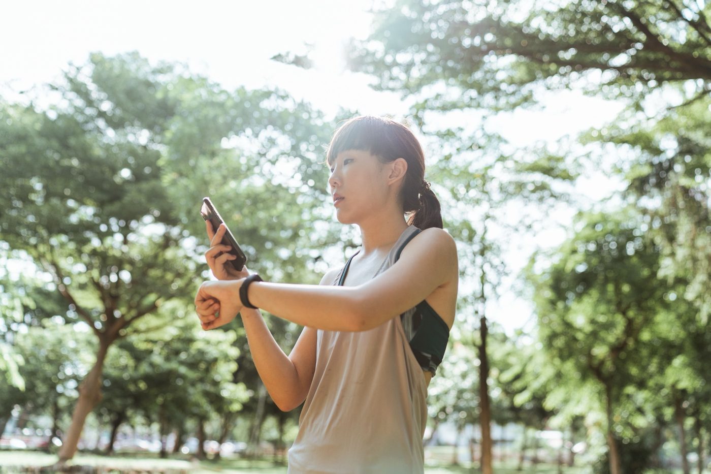 Woman in a sunny park checking her phone and wristband during a workout
